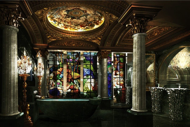 Guests bathe under art covered ceilings lit by standing candelabra. A rain shower and electric bidet toilet are hidden behind a floor to ceiling stained-glass façade.