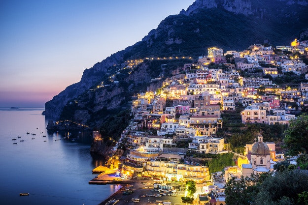 The sun sets over the town of Positano on Italy's Amalfi Coast. We see the boats on the Tyrrhenian Sea and the cliffs in the distance as the lights begin to come on in the town of Positano.