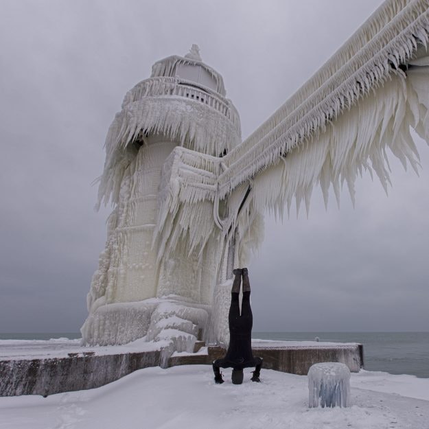 Photographer Joshua Nowicki doing a headstand in a wetsuit beside the frozen lighthouse.