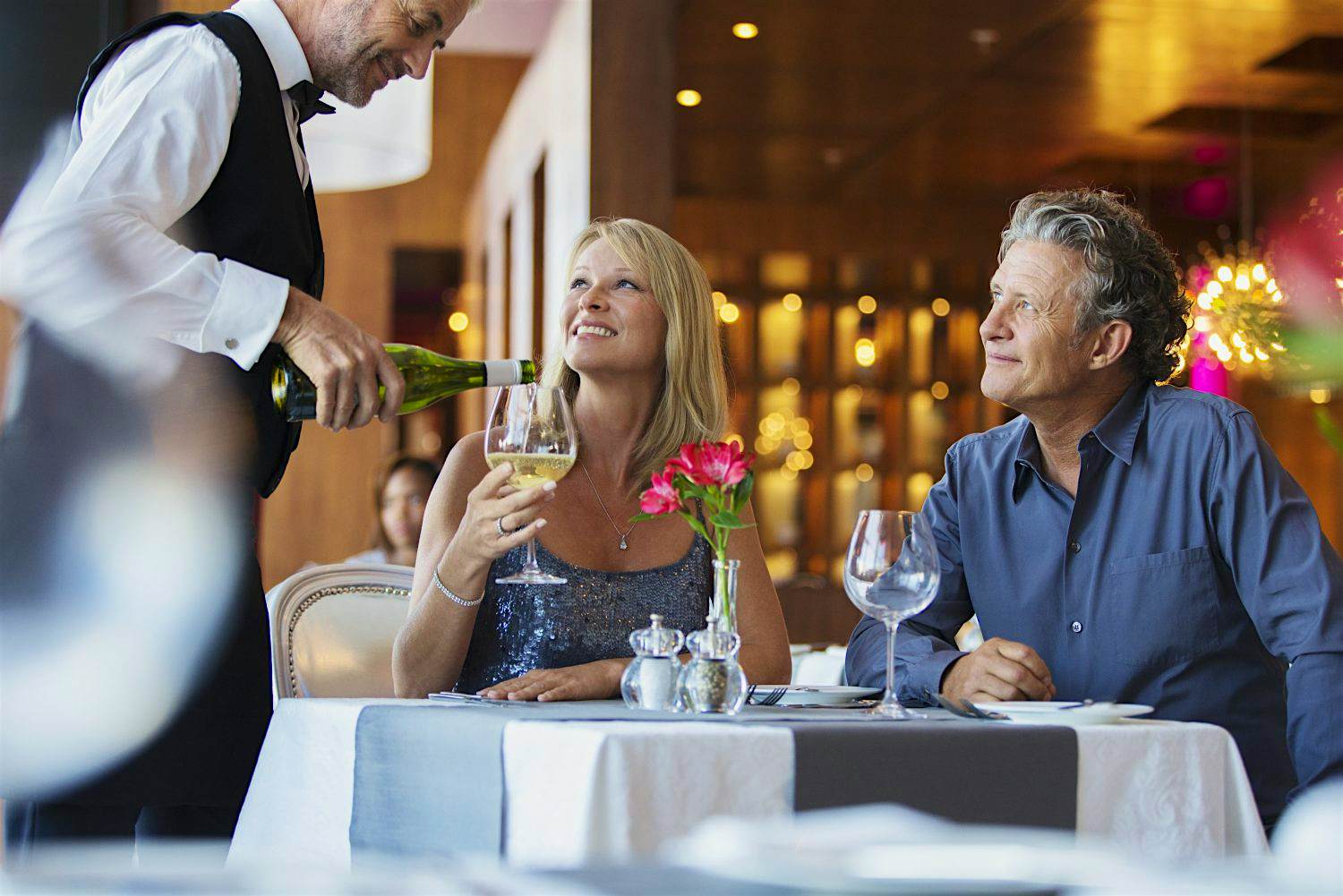 Travel News - Mature couple sitting at restaurant table, waiter pouring white wine into womans glass Travel News - Mature couple sitting at restaurant table, waiter pouring white wine into womans glass