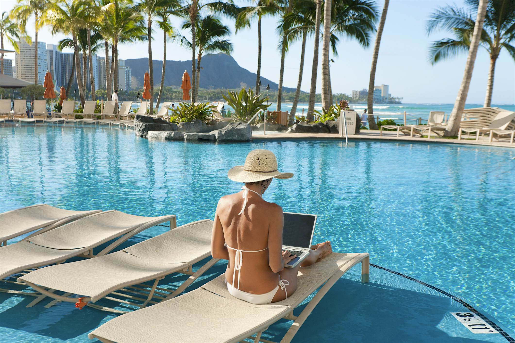 Travel News - Hawaii, Oahu, Waikiki, Woman Lounging At Hotel Poolside With Laptop Computer, Diamond Head In Distance. Travel News - Hawaii, Oahu, Waikiki, Woman Lounging At Hotel Poolside With Laptop Computer, Diamond Head In Distance.