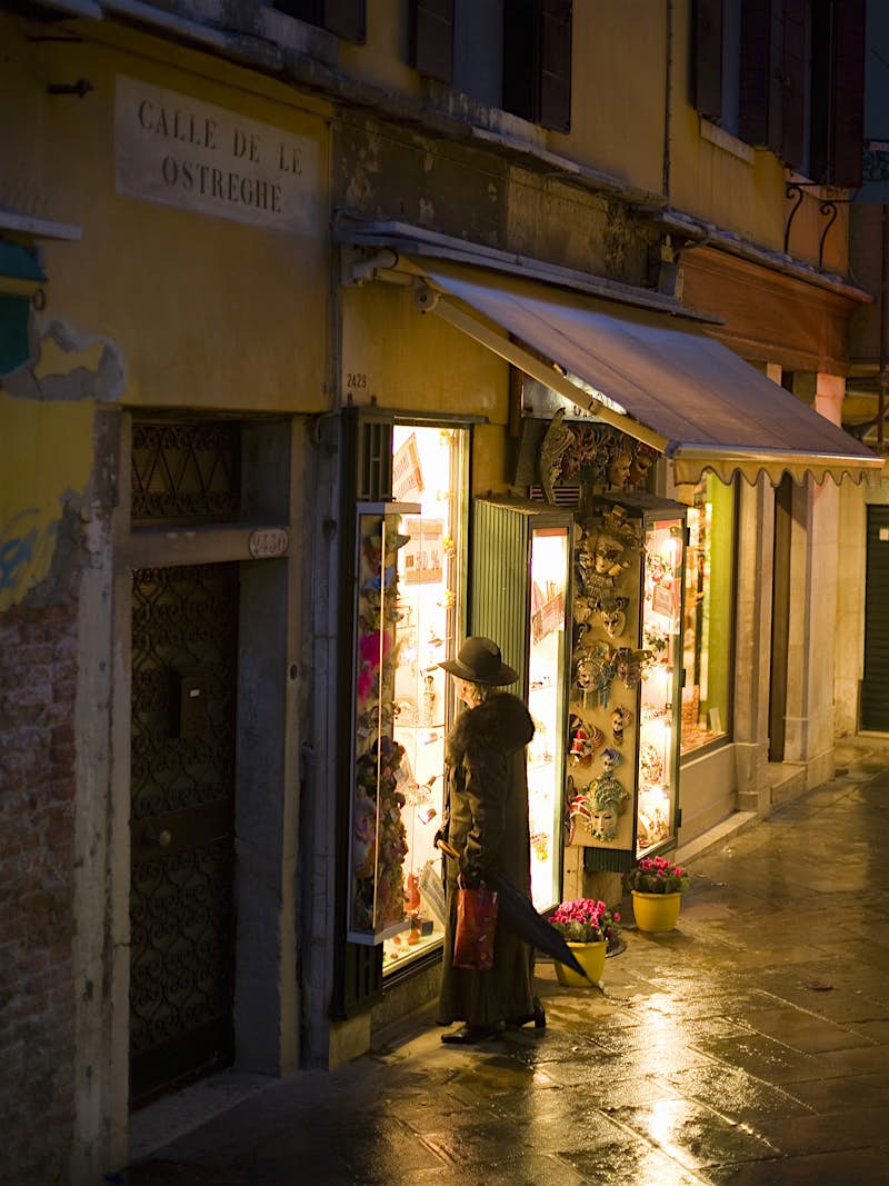 Travel News - A quiet street at night. One person looking into the lighted window of a shop or cafe. Venice offers much more than just palaces and canals. Image by Mint Images