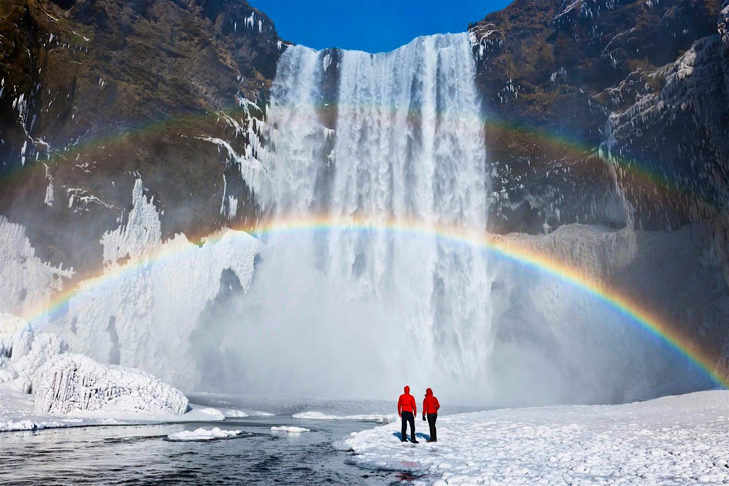 Travel News - Couple under a rainbow by a large waterfall, winter. Skógafoss, Iceland. A rainbow by a large waterfall in Skógafoss, Iceland. Image by Jeremy Walker/Getty Images