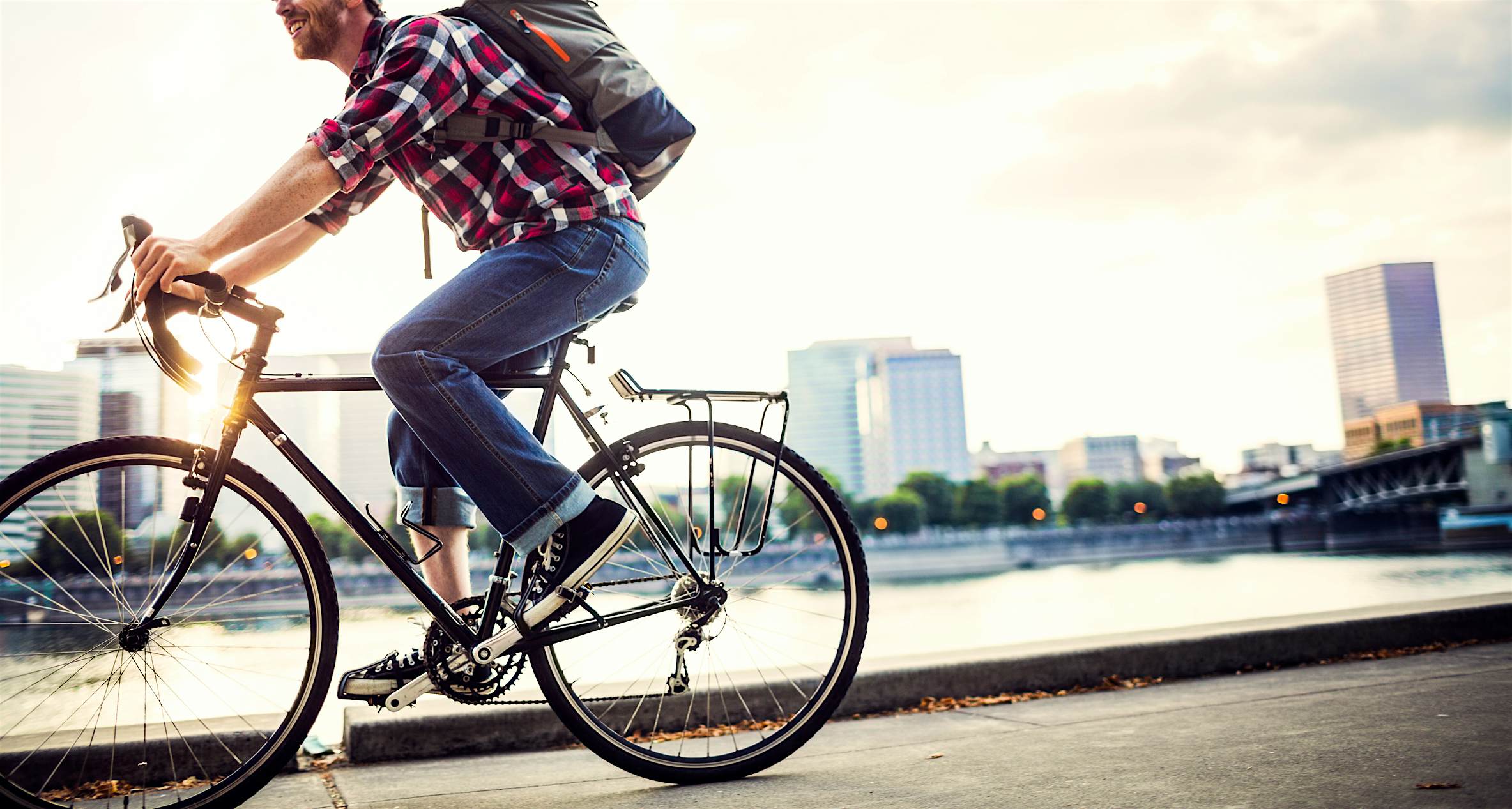 Travel News - Bike Commuter in Portland Oregon A young man commuting in an urban city environment on his street bicycle, a waterproof bike bag on his back. This is the Eastbank Esplanade in Portland, Oregon, that follows along the Willamette river. Downtown is visible across the water, the sun shining brightly between the buildings. Horizontal, in-camera lens flare, in motion.