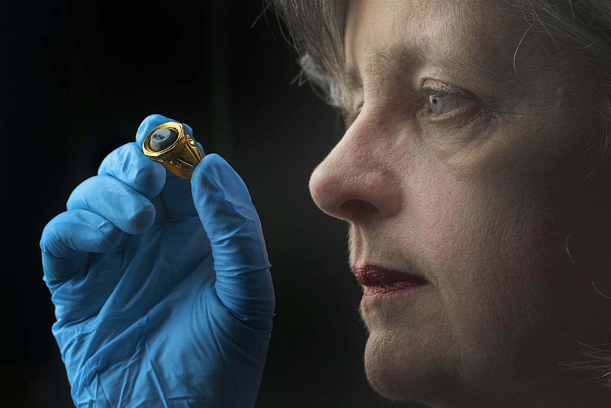 Travel News - Curator Jackie Keily looks closely at a gold ring from a woman's burial, which depicts a scene from Horace's Satires © Museum of London Curator examines a gold ring