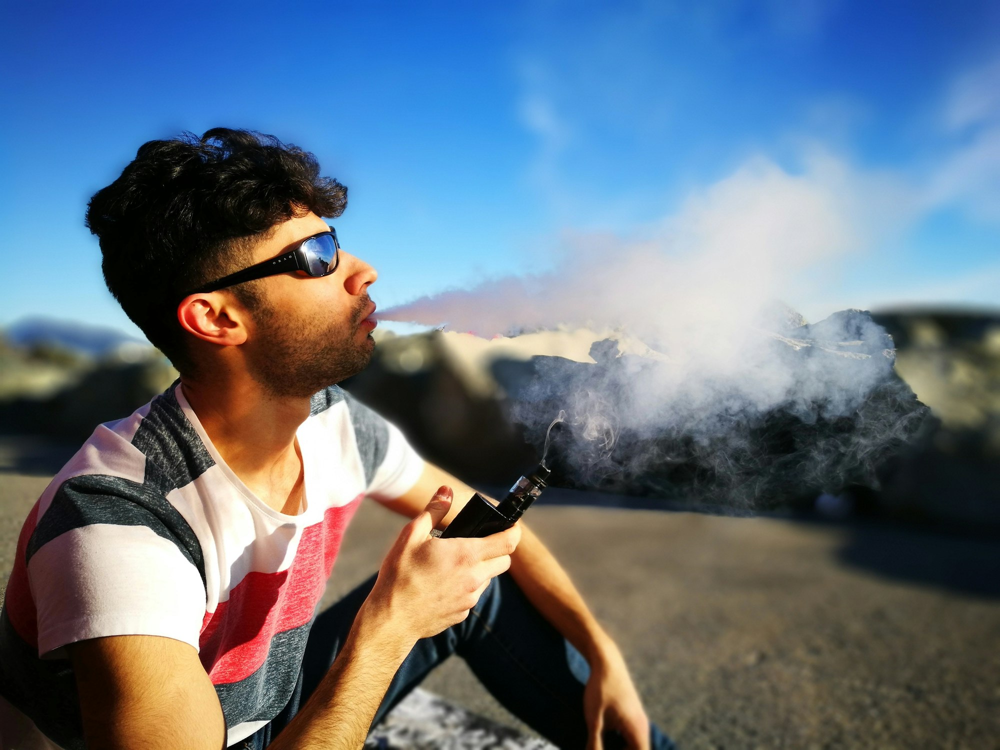 Travel News - Young Man Smoking Electronic Cigarette While Sitting On Field Against Sky