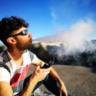 Travel News - Young Man Smoking Electronic Cigarette While Sitting On Field Against Sky