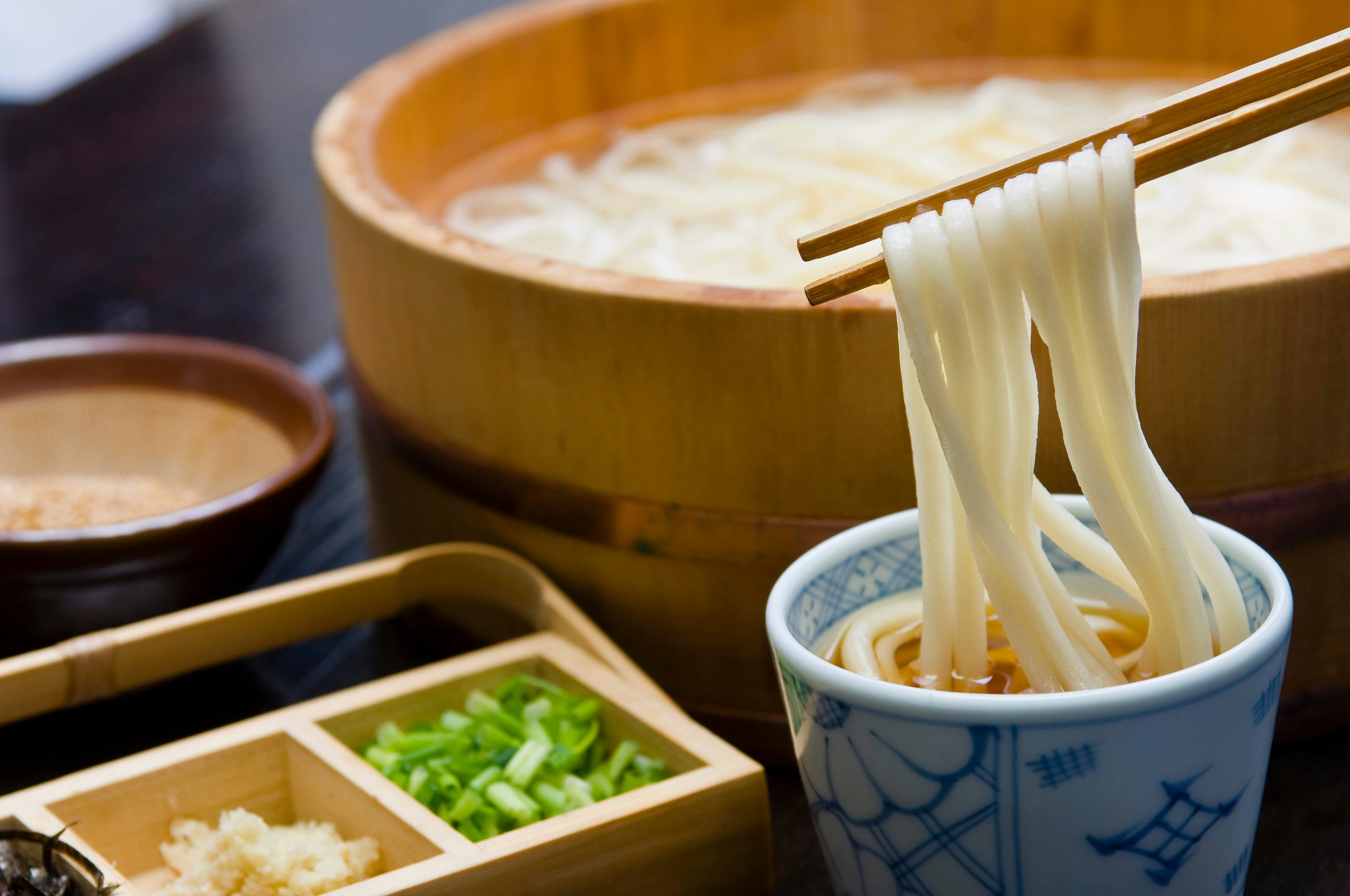 A pair of wooden chopsticks emerge from the right side of the frame to dunk several strands of white udon noodles into a small cup of broth. In the background is a large wooden tub of udon noodles, while to the left sits a wooden box filled with green onions in one compartment and ginger in the other.