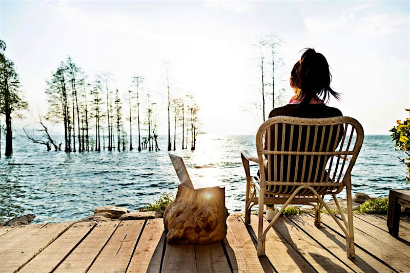 Travel News - Young woman working on her laptop while sitting by the lake. A woman working on her laptop while sitting by the lake.