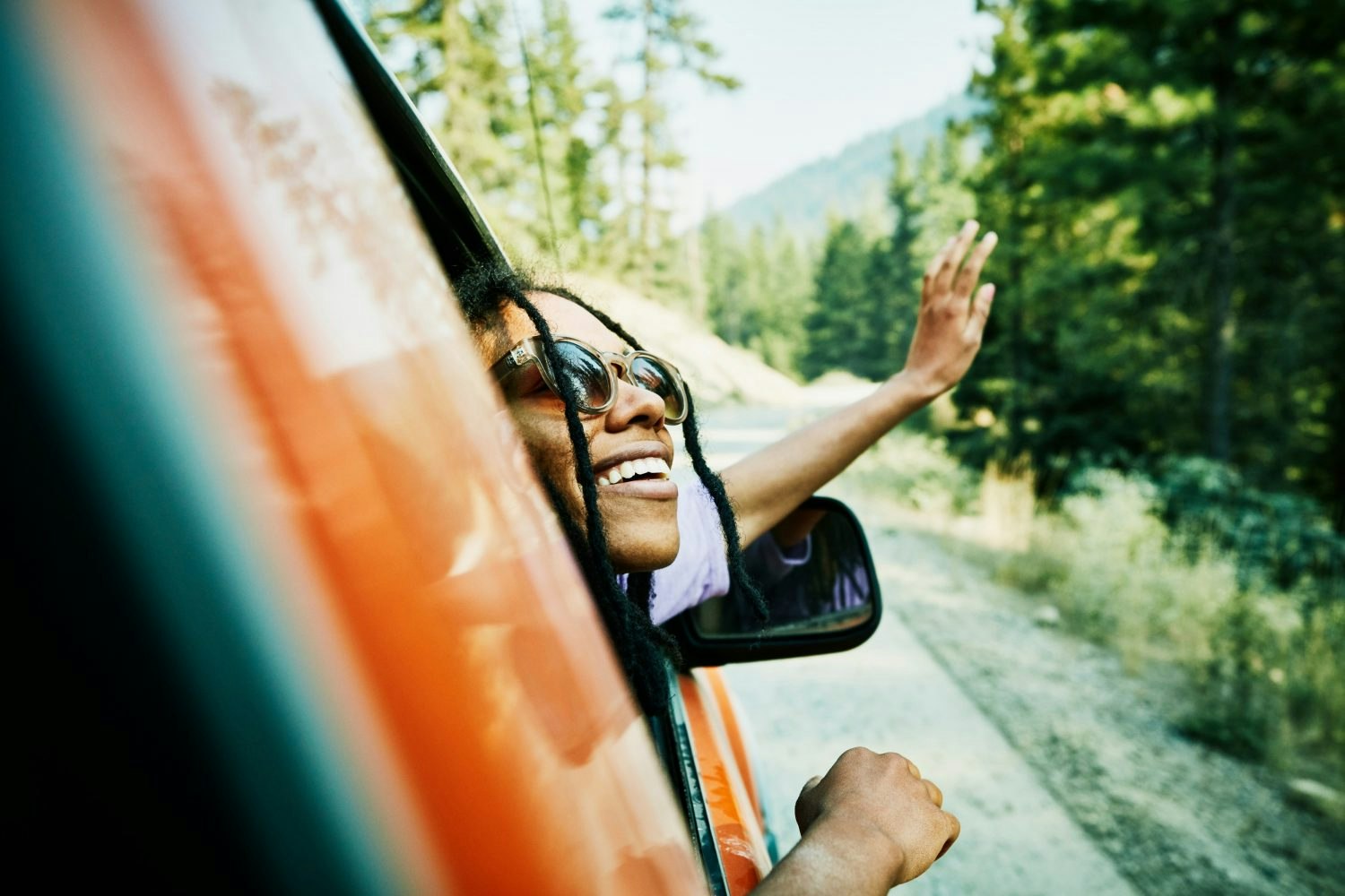 Smiling woman with head and hand out of car window enjoying view of mountains