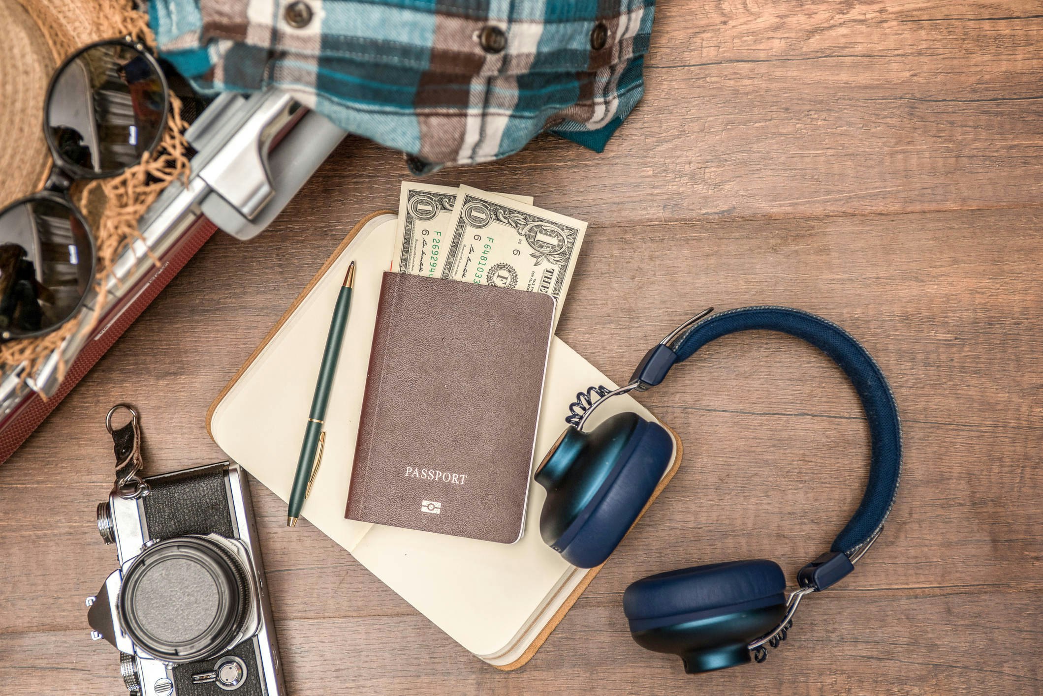 A table covered with items: a camera, passport with two US dollars sticking out, a notebook and a pen, sunglasses, and headphones
